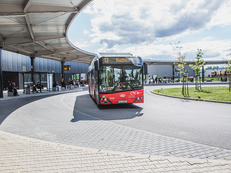 Fotografię wykonano na dworcu autobusowym. Widoczna jest długa łukowata wiata przystankowa i pasażerowie czekający na transport. Na pierwszym planie znajduje się czerwony autobus z wyświetloną informacją: 13 Kuźnia. Zdjęcie pochodzi z archiwum beneficjenta.