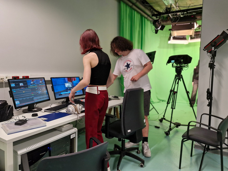 Na zdjęciu widzimy studio filmowe. Z prawej widoczne jest zielone pomieszczenie do wykonywania zdjęć. Na pierwszym planie – stanowiska komputerowe obsługiwane przez dwójkę uczniów. Fotografia pochodzi z archiwum beneficjenta.