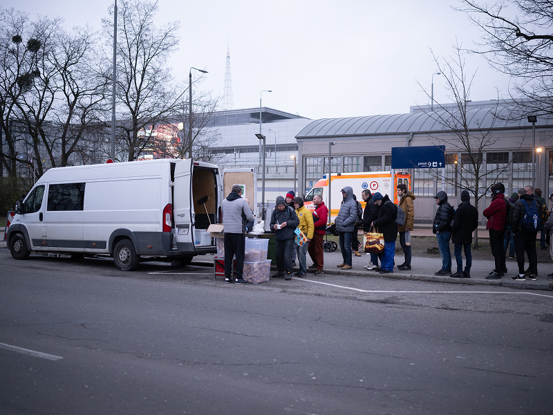 Na zdjęciu widoczna jest grupa bezdomnych, która stoi w kolejce po darmowy, ciepły posiłek. Wydawany jest z samochodu dostawczego. Autorem fotografii jest Maciej Motylewski.