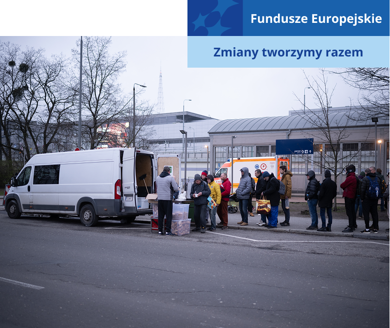 Na zdjęciu widoczna jest grupa bezdomnych, która stoi w kolejce po darmowy, ciepły posiłek. Wydawany jest z samochodu dostawczego. Autorem fotografii jest Maciej Motylewski.